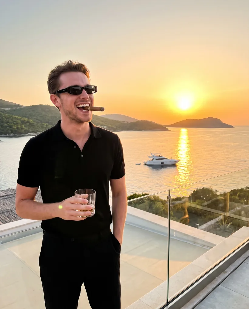 A man laughing on a rooftop terrace at sunset, whisky in hand, a yacht on the bay below