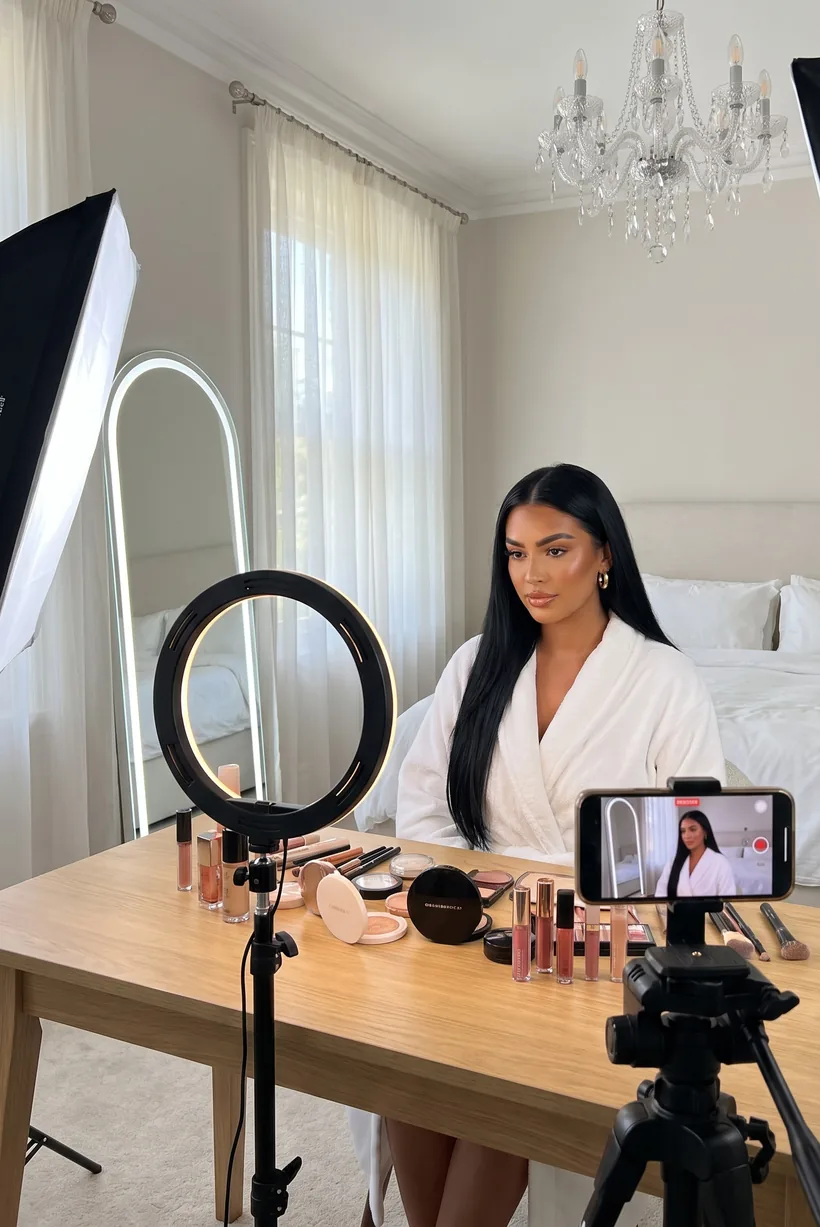 A beauty creator in a white robe at a makeup table, a ring light and a phone recording her