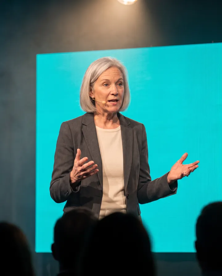 A speaker mid-gesture on a stage with a teal backdrop